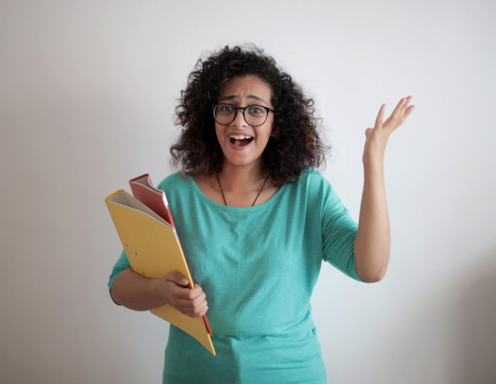 Woman in glasses with curly hair wearing teal sweater holding yellow and red folders in one hand and holding up the other hand.